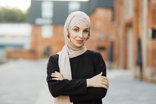Portrait of young muslim woman wearing hijab head scarf in city while looking at camera. Closeup face of cheerful woman covered with headscarf smiling outdoor. Casual islamic girl in town.