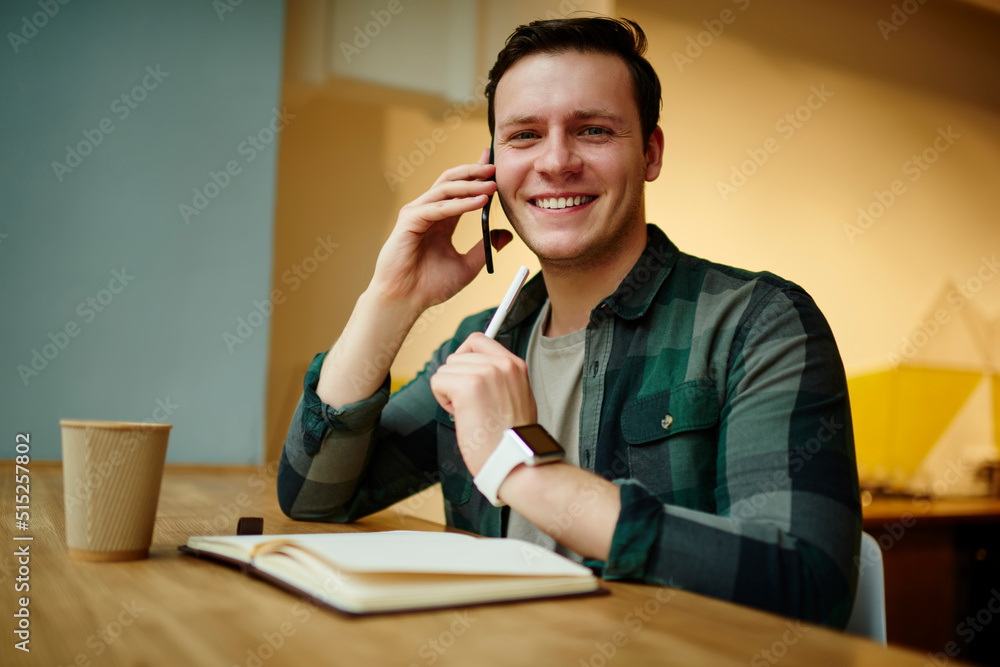 Poster portrait of young smiling man calling via mobile phone while sitting at the desk with netbook. worki