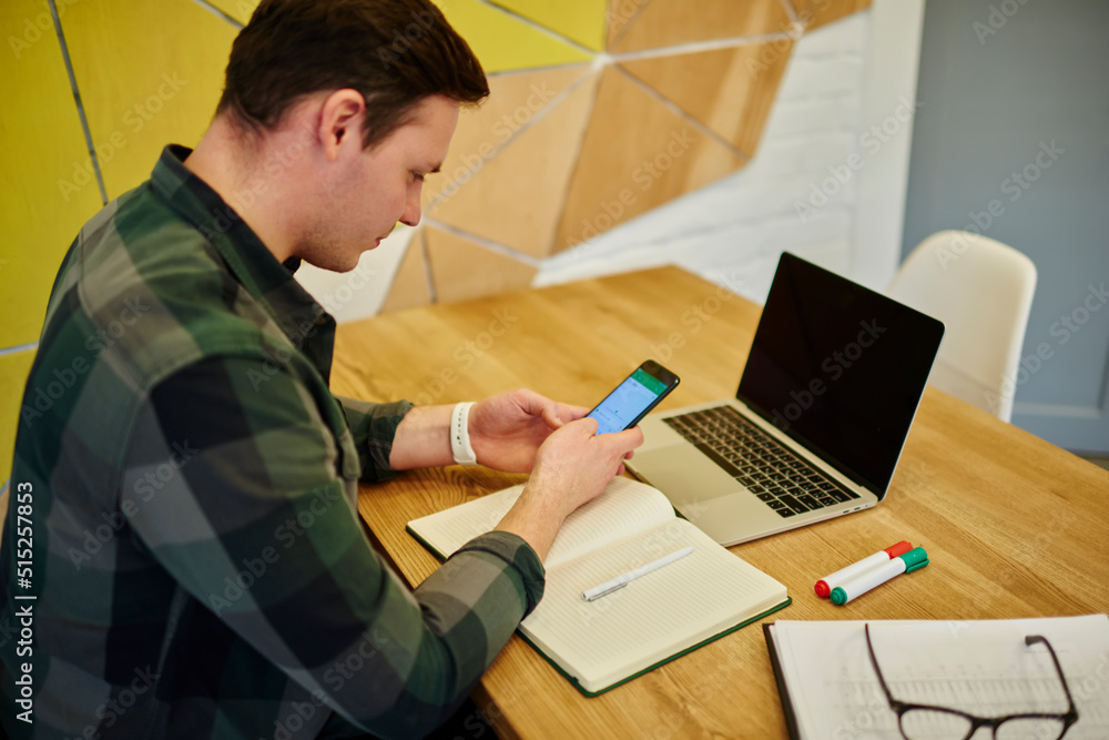 Poster young thoughtful man texting message on mobile phone while sitting at the desk with laptop computer 
