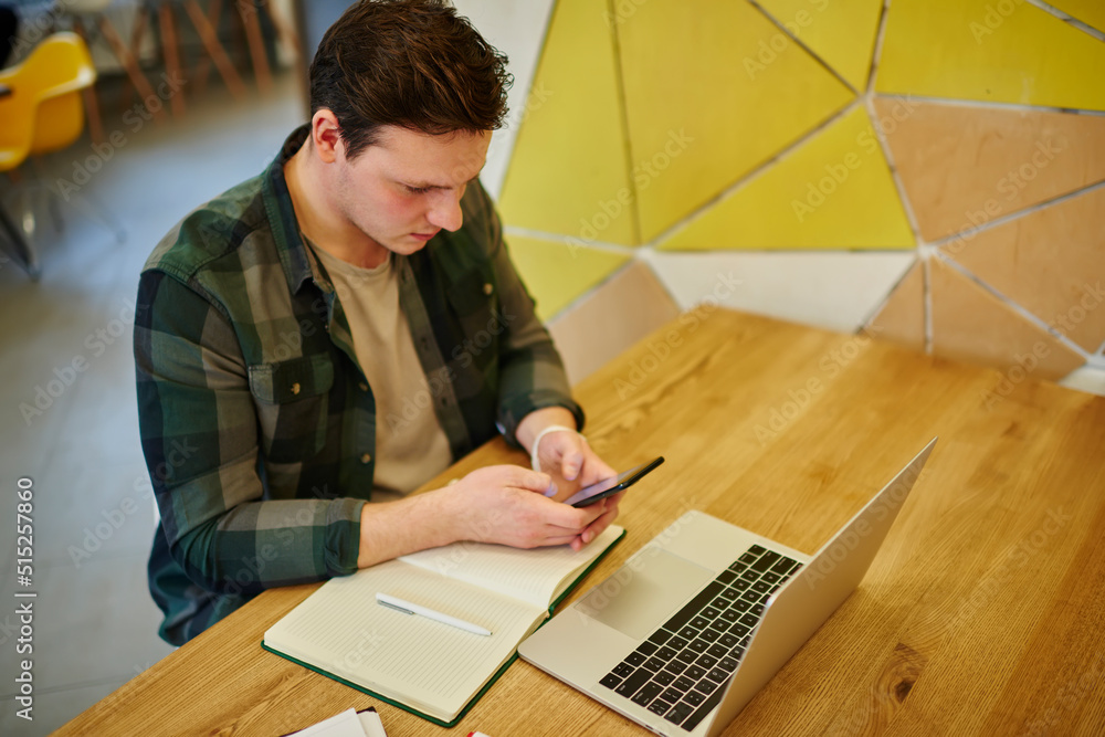 Poster young thoughtful man texting message on mobile phone while sitting at the desk with laptop computer 