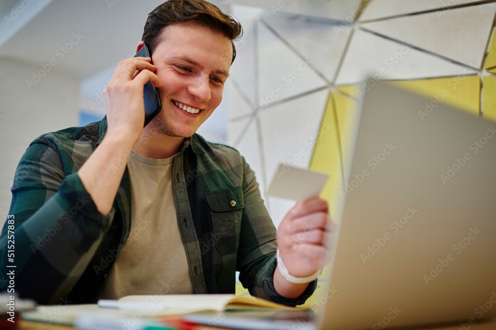 Poster young smiling man calling via mobile phone while sitting at the desk with laptop computer. working o