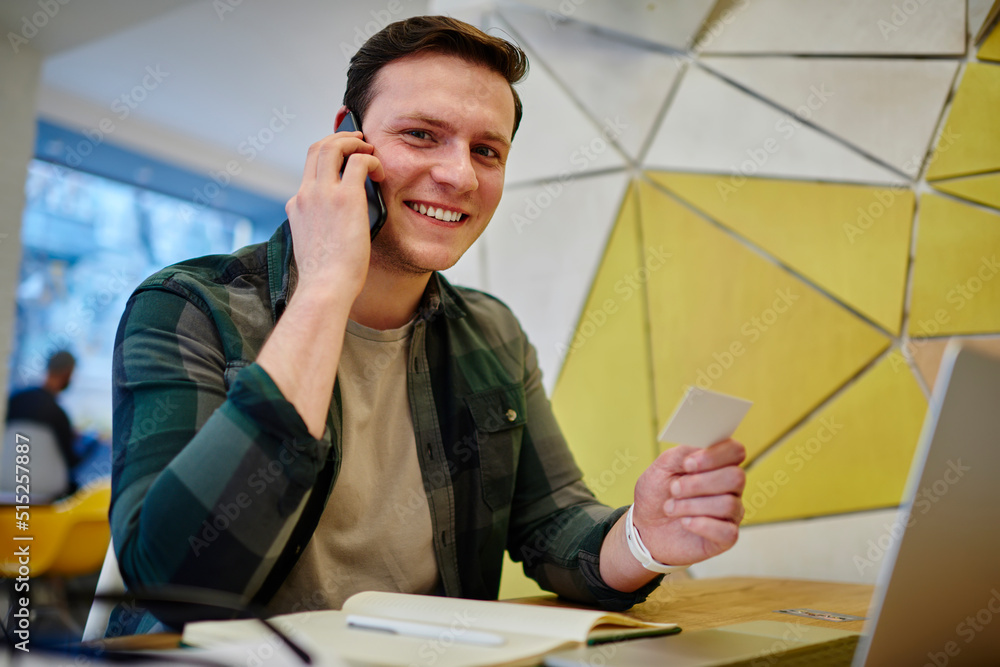 Poster young smiling man calling via mobile phone while sitting at the desk with laptop computer. working o