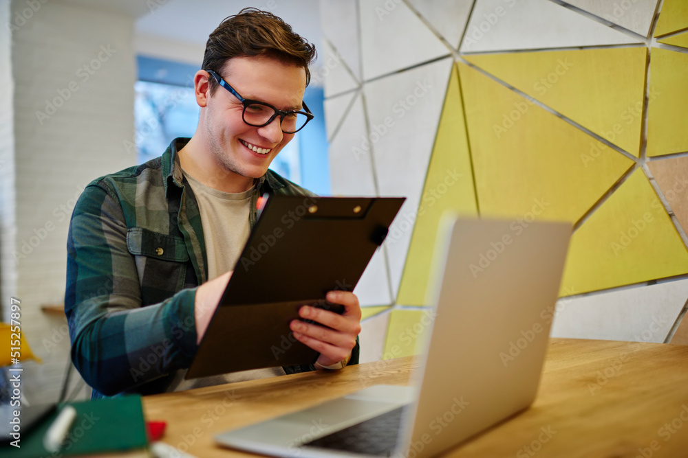 Poster young smiling male student writing notes in notebook while learning online on laptop computer. man f