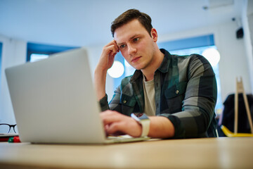 Young thoughtful man sitting front laptop computer while working online on freelance. Male student learning via netbook. Reading article on website