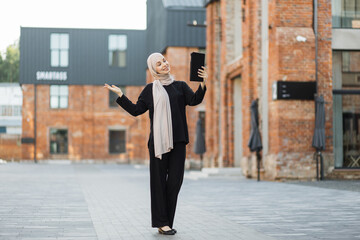 Full lens portrait young Muslim Arab woman in hijab takes a selfie of herself with the city background. Elegant, tall, slim and wearing black clothes and turban, smiling while takes selfie on tablet.