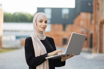 Attractive smiling muslim woman in hijab texting on notebook standing on background of buildings in city street. Portrait of young arabian businesswoman using laptop in city.