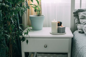 Bedside table with burning modern candle, home plant. Hygge, cozy concept.