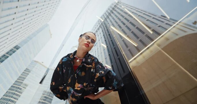 Wide angle POV urban style seductive girl wearing colored jacket cool futuristic glasses bends over and looks into camera near high skyscraper modern building. Portrait handsome female fashion model