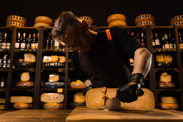 Handsome man worker of cheese shop cut limited gouda with knife. Cheese sommelier working in cheese...