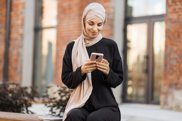 Pretty smiling muslim business woman in hijab sitting on bench and using smart phone during video...