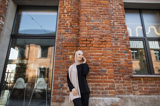 Cheerful young islamic businesswoman talking on mobile phone while standing near red brick wall. Beautiful young woman with hijab working using smartphone during break. - Powered by Adobe
