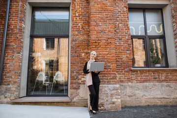 Pretty smiling muslim business woman in hijab standing near office centre and using laptop during video chat. Happy arabic girl standing outdoor near work office and checking email on digital gadget.