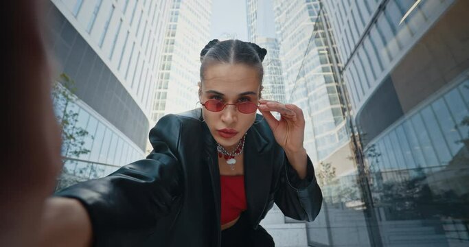 POV portrait of stylish serious woman, seductive trendy urban style female touching cool red eye glasses taking selfie with smartphone looking at camera standing outdoors overlooking glass skyscrapers