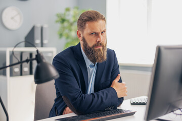 Fototapeta premium Pensive mature man in stylish business wear sitting at desk with modern pc. Successful caucasian male person working at bright office. Career and people concept.