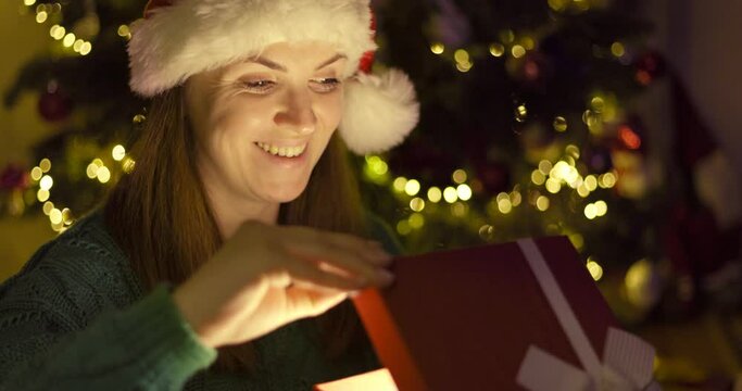 Beautiful young woman in a Christmas hat opening Christmas gift box with excited surprised face feeling happy on New Year's Eve sitting at home near glowing christmas tree. Magic moments.