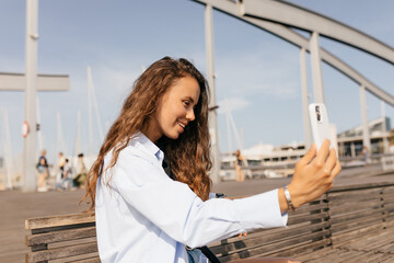 Pretty charming lady with long loose wavy hair in blue shirt making selfie on the pier in sunny warm day. Lovely girl walking outdoor 