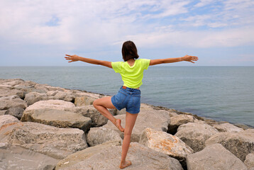slender girl on the rocks of the dike near the sea while performing gymnastic exercises to keep balance with one leg