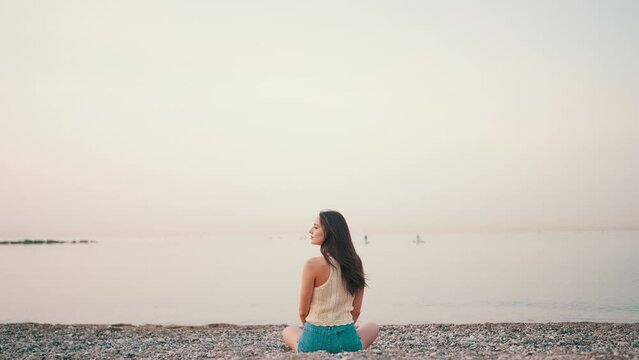 Beautiful brown-haired woman with long hair sits on the beach on seascape background. Gorgeous girl wearing top and denim shorts. Slow motion. Back view
