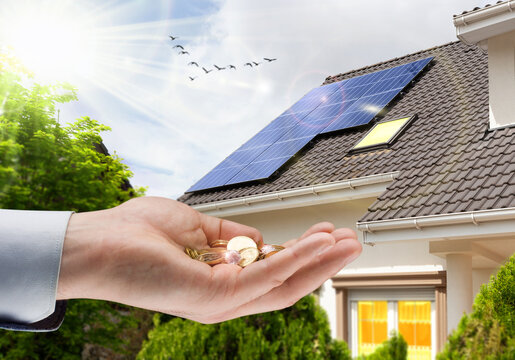 Hand with money against background of house with installed solar panels. Renewable energy and saving money. Conceptual image