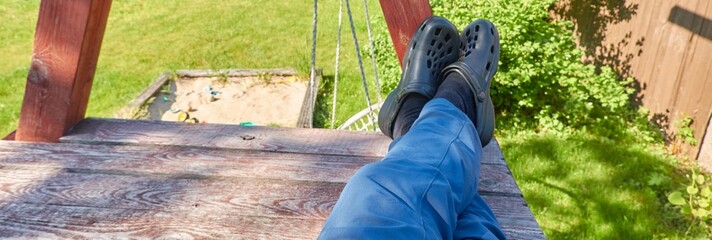 Man resting on wooden swing bench. Foam clogs, blue jeans. Backyard, garden, playground. Sunny day....