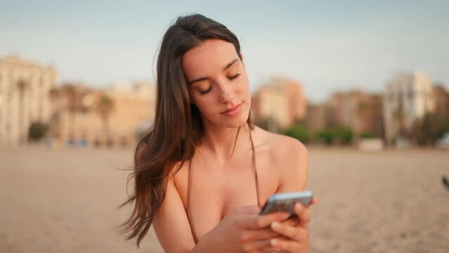 Slim beautiful brown-haired woman with long hair is using mobile phone sitting on the beach. Gorgeous girl scrolling information into smartphone. Camera moves sideway to the right side