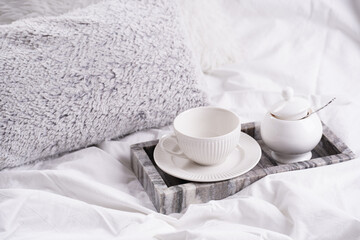 Fototapeta na wymiar an empty white porcelain cup for coffee or tea and saucer with sugar in grey marble tray on white bed sheets among fluffy pillows - breakfast in bed
