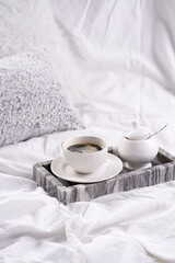 Fototapeta na wymiar a white porcelain cup with black coffee americano and saucer with sugar in grey marble tray on white bed sheets among fluffy pillows - breakfast in bed