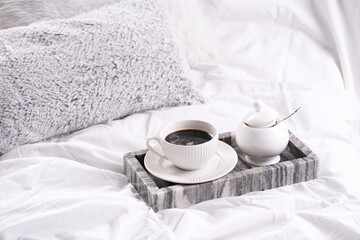a white porcelain cup with black coffee americano and saucer with sugar in grey marble tray on white bed sheets among fluffy pillows - breakfast in bed