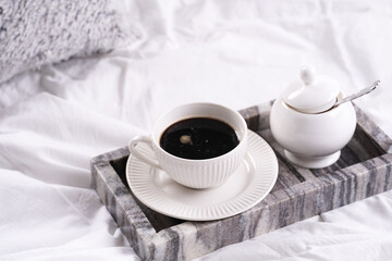 Fototapeta na wymiar a white porcelain cup with black coffee americano and saucer with sugar in grey marble tray on white bed sheets among fluffy pillows - breakfast in bed