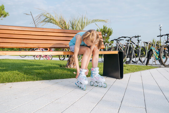 A girl in roller skates fastens a roller lock in a park on a bench.