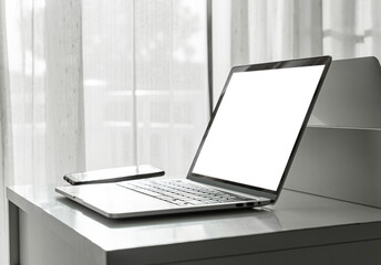 Computer laptop with blank screen and smartphone on white table in home interior or office background.