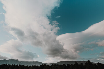 Hills with forest line on horizon under blue sky. Mountain range under cumulus clouds. Approach of rain front, deterioration of weather. Rays of sun break through dense layers of atmosphere. 