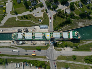 A direct overhead, top-down aerial view of a bulk carrier cargo ship passing through a tight urban canal at a drawbridge crossing during the day.