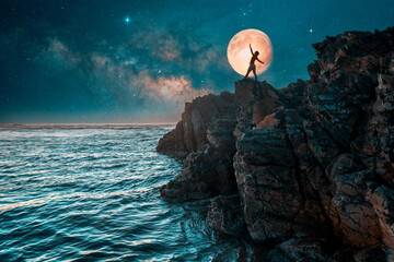 silhouette of a woman with open arms dancing on the cliff over the ocean with full moon in the...