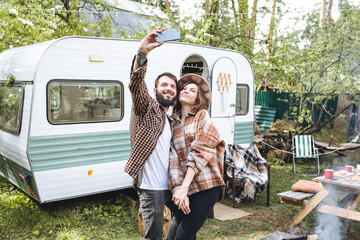 Young beautiful loving couple travelling across country in the van. Millennial man and woman in a travel camper. Cozy atmosphere, vacations vibe. Playing guitar, drinking tea, taking selfie photo - obrazy, fototapety, plakaty