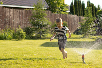 A little wet boy runs barefoot on the lawn next to the sprinkler. The concept of a happy, carefree...