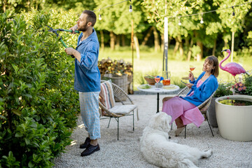 Young stylish family spend summer time at backyard of their country house on nature. Man pruning...