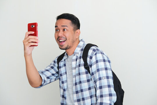 Adult Asian man wearing backpack showing excited expression when taking a photo using handphone