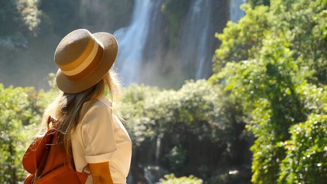 Travel woman enjoy amazing view of waterfall nature. Female traveler in hat with backpack in Asia adventure or journey. Cinematic video of girl in tropical vacation.