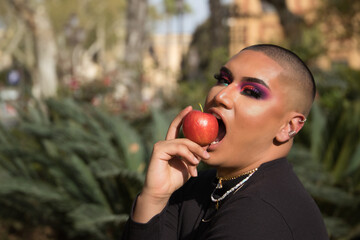 Portrait of non-binary person, young and South American, heavily make up, biting a red apple that...