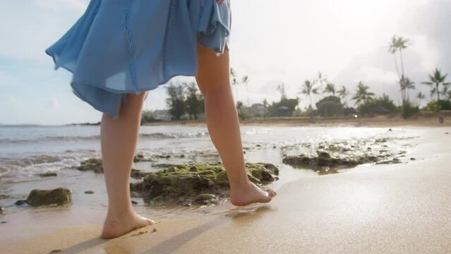RED camera Slow Motion golden sunrise light highlighting nature. Slim female legs of nature background. Close up woman in flattering blue beach dress walking barefoot by sandy beach in scenic seascape
