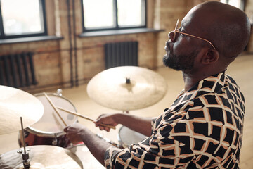 Young African musician in eyeglasses playing drums at drum kit sitting at studio