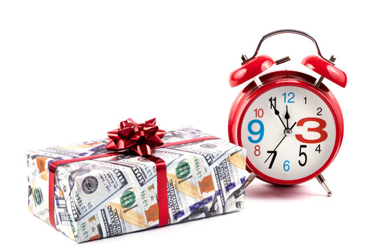 Red alarm clock and a gift box wrapped in paper with a picture of 100 american dollars bills on a white background
