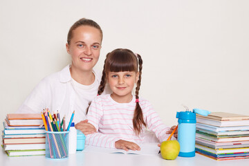 Horizontal shot of smiling female sitting with her daughter at the desk isolated over white background, doing homework together, posing surrounded with books, looking at camera with smiles.