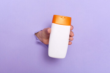 Closeup of person's hand holding cosmetics bottle or shampoo in the hole of purple paper wall, copy space for your design, advertisement ara for promotional text. Banner sales.