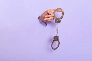 Person's hand holding handcuffs in purple paper hole, silver shackle in human's hand breaking through lilac background. Criminality, misdemeanor.