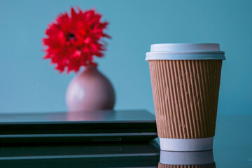 Cup of coffee to go on a table in focus. Closed note book computer and red flower out of focus. Blue color background. Simple office space concept