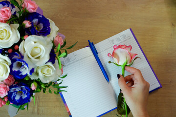 Flatlay. Female hands hold a rose flower. Notebook, diary with a pen on a wooden table near a...