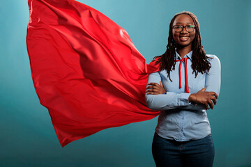Mighty looking tough superhero woman wearing red hero costume cape standing with arms crossed on blue background. Portrait of young adult person posing as justice defender while smiling at camera.