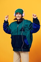 a happy, joyful woman in a green jacket and hat enjoys music while standing in pink headphones on a yellow background dancing. Vertical photography